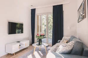 A modern living room with a flat-screen TV, a white console, a round coffee table with a vase of flowers, a grey sofa with a fluffy throw, and a large window with curtains. - Puszcza Białowieska Apartments by 2HomeRent (Nieznany Bór)