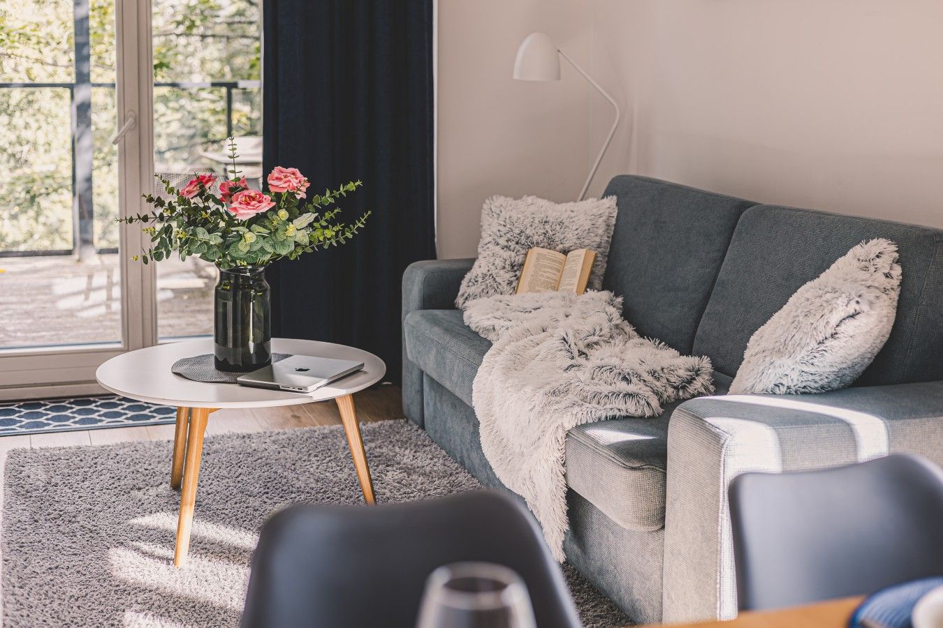 A modern living room with a grey sofa, a white side table, a vase with flowers, and a book on the sofa.