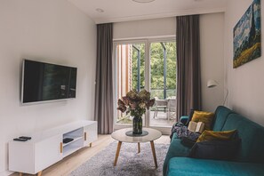 A modern living room with a flat-screen TV, a white entertainment unit, a teal sofa with cushions, a round coffee table with a vase of flowers, and a large window with curtains. - Puszcza Białowieska Apartments by 2HomeRent (Nieznany Bór)