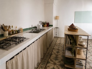 A kitchen with a gas stove, sink, and a long countertop. There is a shelf with kitchen utensils and a lamp. - An authentic Salento home with a private courtyard, in the heart of Salento (Giuliano Di Lecce)