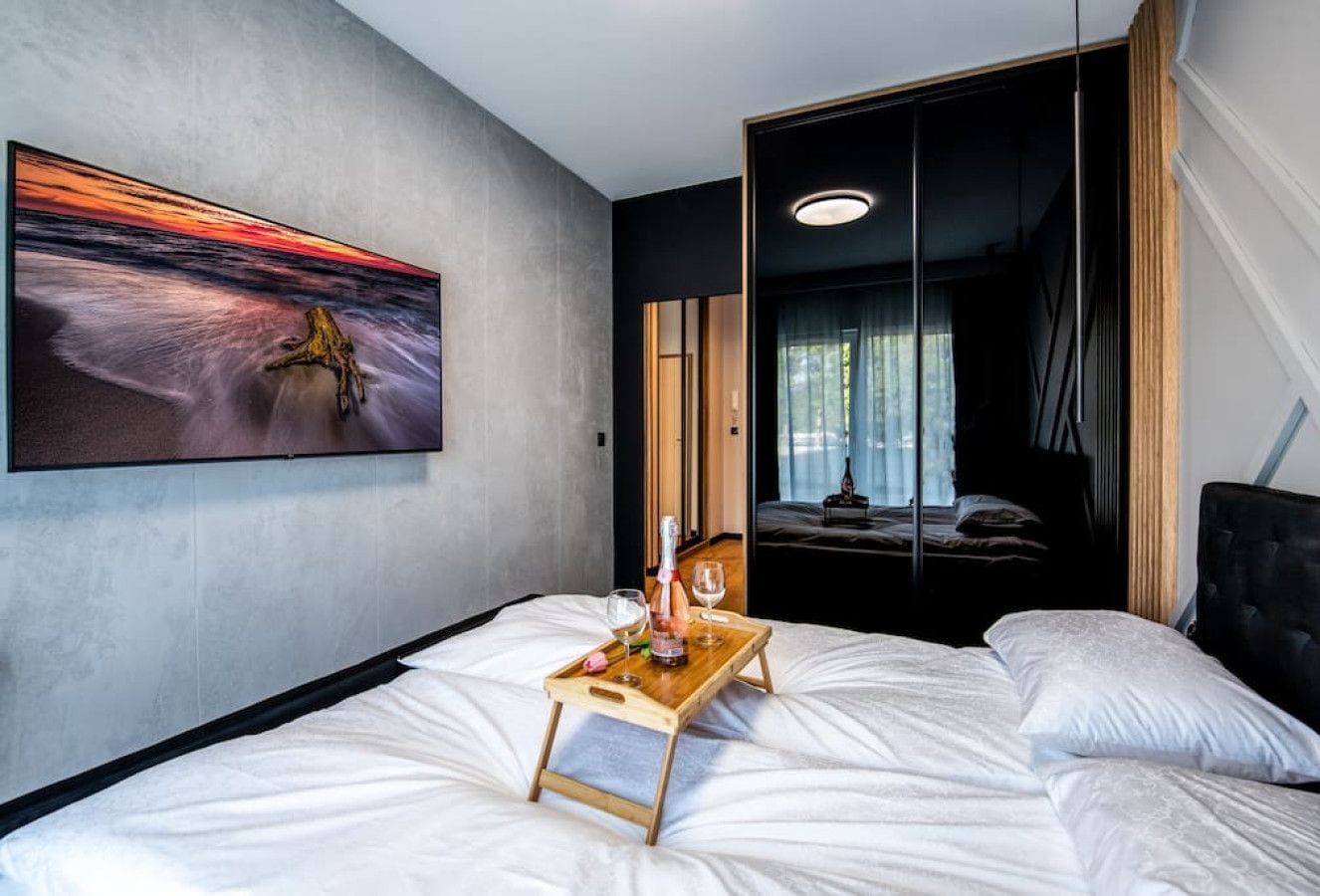 A modern hotel room with a bed, a bedside table with a tray holding a bottle and glasses, a large painting on the wall, and a view of another room through a sliding door.