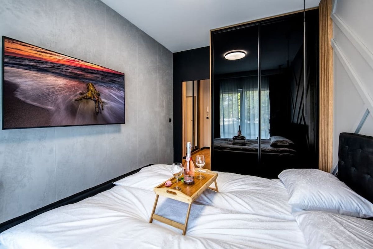 A modern hotel room with a bed, a bedside table with a tray holding a bottle and glasses, a large painting on the wall, and a view of another room through a sliding door.