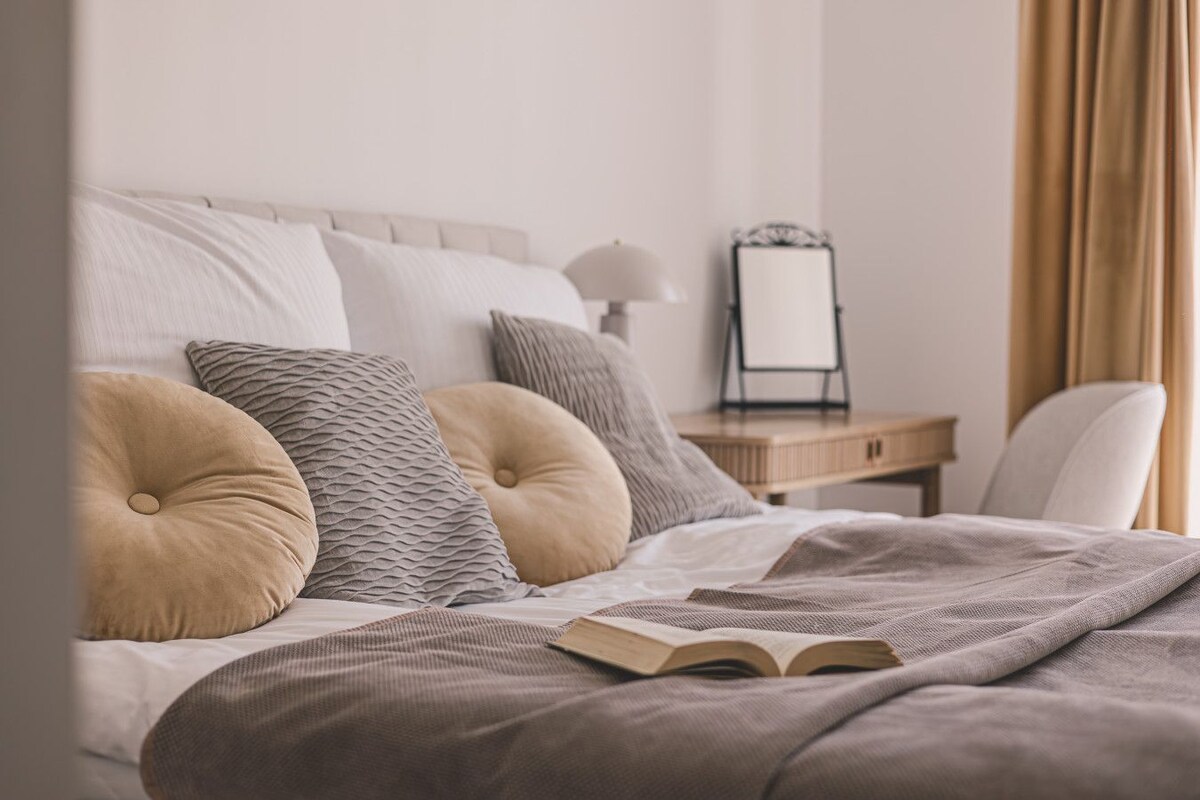 A neatly made bed with a book, a bedside table with a lamp, and a mirror.