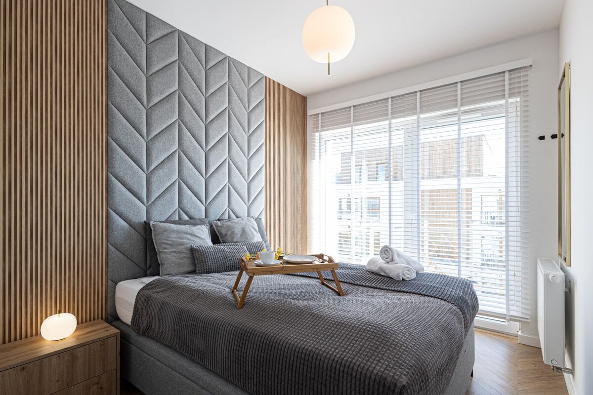 A modern bedroom with a large bed, a bedside table with a lamp, a tray with a cup and saucer, and a window with blinds.