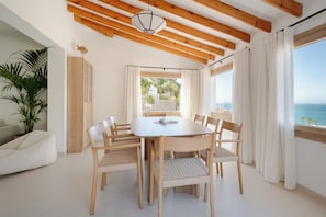 A dining area with a round table surrounded by chairs, wooden beams on the ceiling, a potted plant, and a view of the sea through a window. - Exclusive Villa Pharos (Almuñécar)