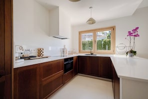 A modern kitchen with wooden cabinets, a built-in oven, and a window with a view of greenery. - Exclusive Villa Pharos (Almuñécar)