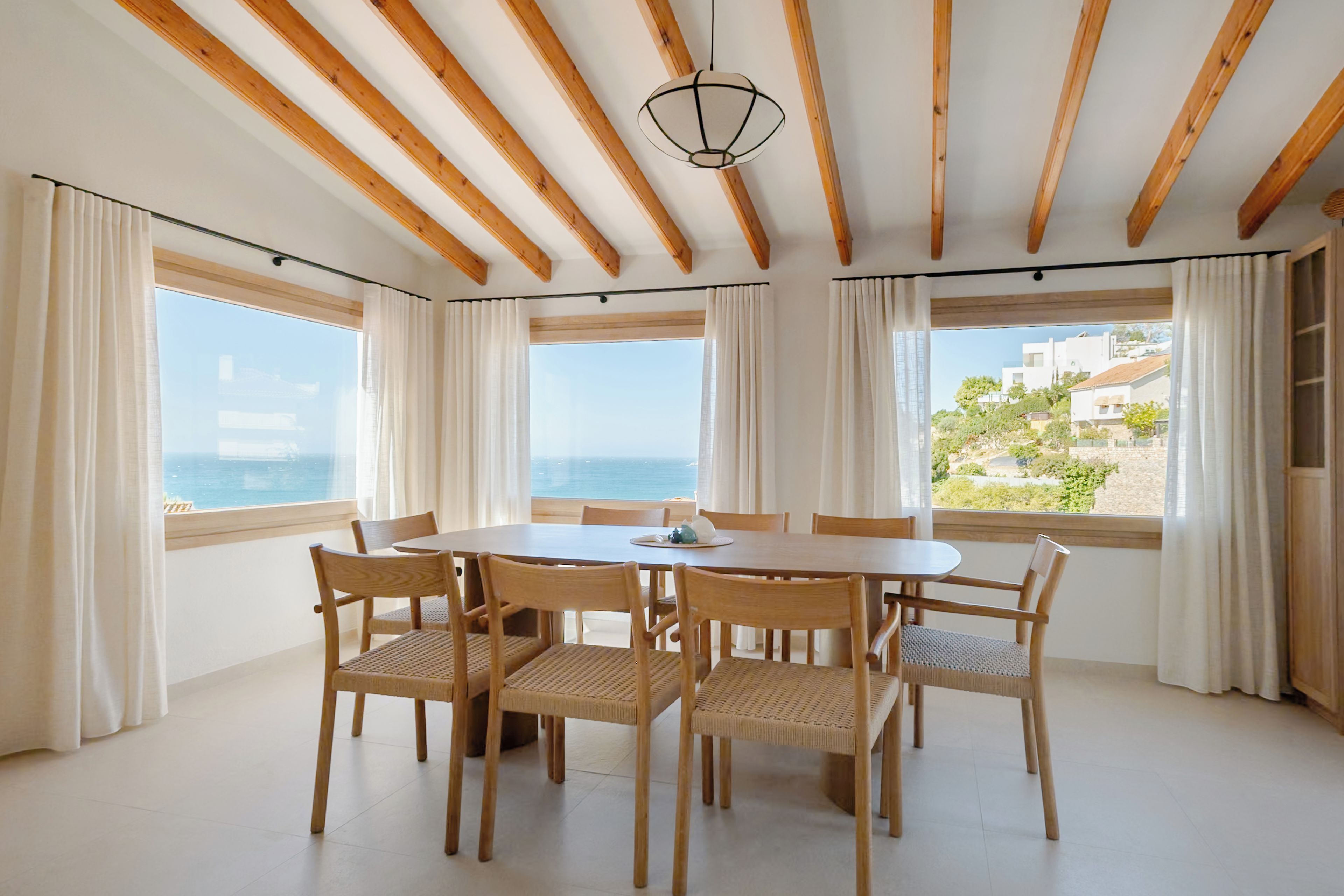 Uma sala de jantar com mesa e cadeiras de madeira, janelas amplas com cortinas e vista para o mar.