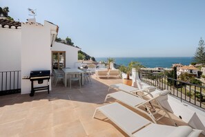 Une terrasse équipée d’un barbecue, d’une table à manger, de chaises et de fauteuils, offrant une vue sur la côte.