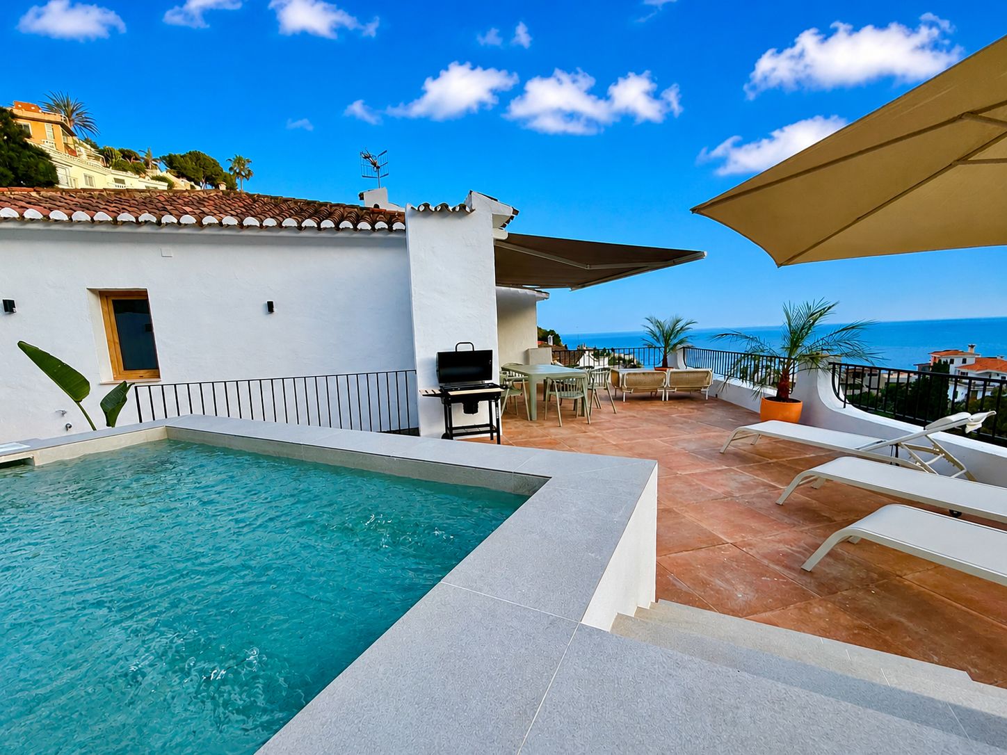 Uma área à beira da piscina com vista para o mar, mobiliário de exterior e um espaço para churrasco.