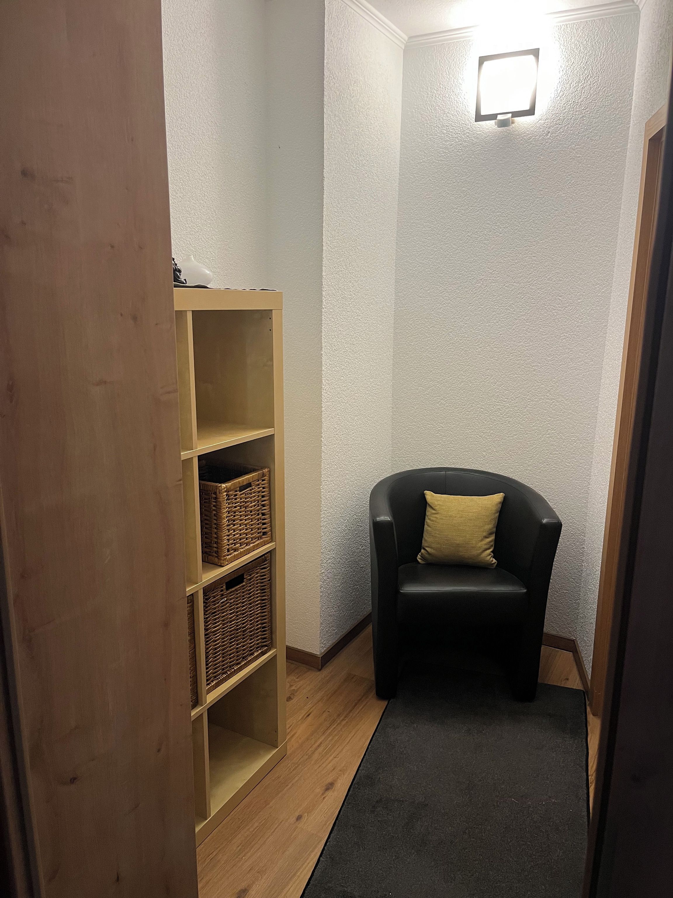 Ein Eckzimmer mit einem schwarzen Ledersessel, einem Holzbodenregal mit Körben und einer strukturierten Wand.