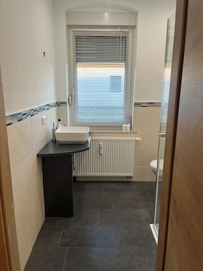 Un baño con lavamanos blanco, encimera negra, ventana con persianas, inodoro y zona de ducha.