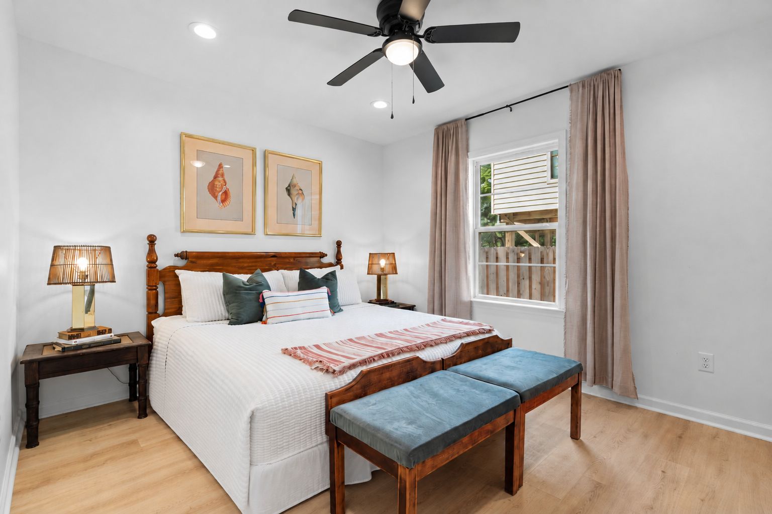 Une chambre à coucher comprenant un lit, des tables de chevet, un ventilateur de plafond, une fenêtre avec des rideaux et des œuvres d’art accrochées au mur.