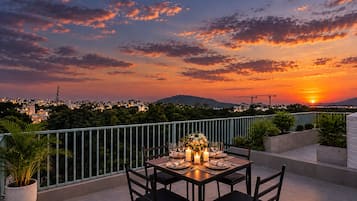 A rooftop terrace with a table set for dining, overlooking a cityscape at sunset.