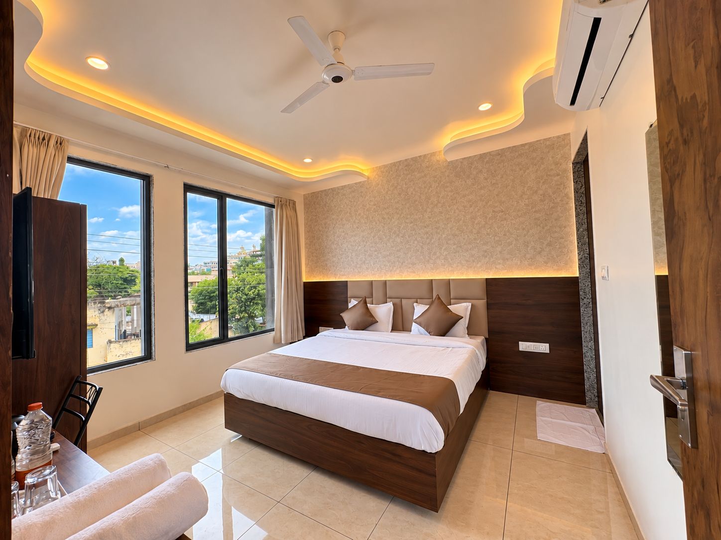 A hotel room with a bed, a ceiling fan, an air conditioning unit, a large window with a view, and a dining area with a table and chairs.