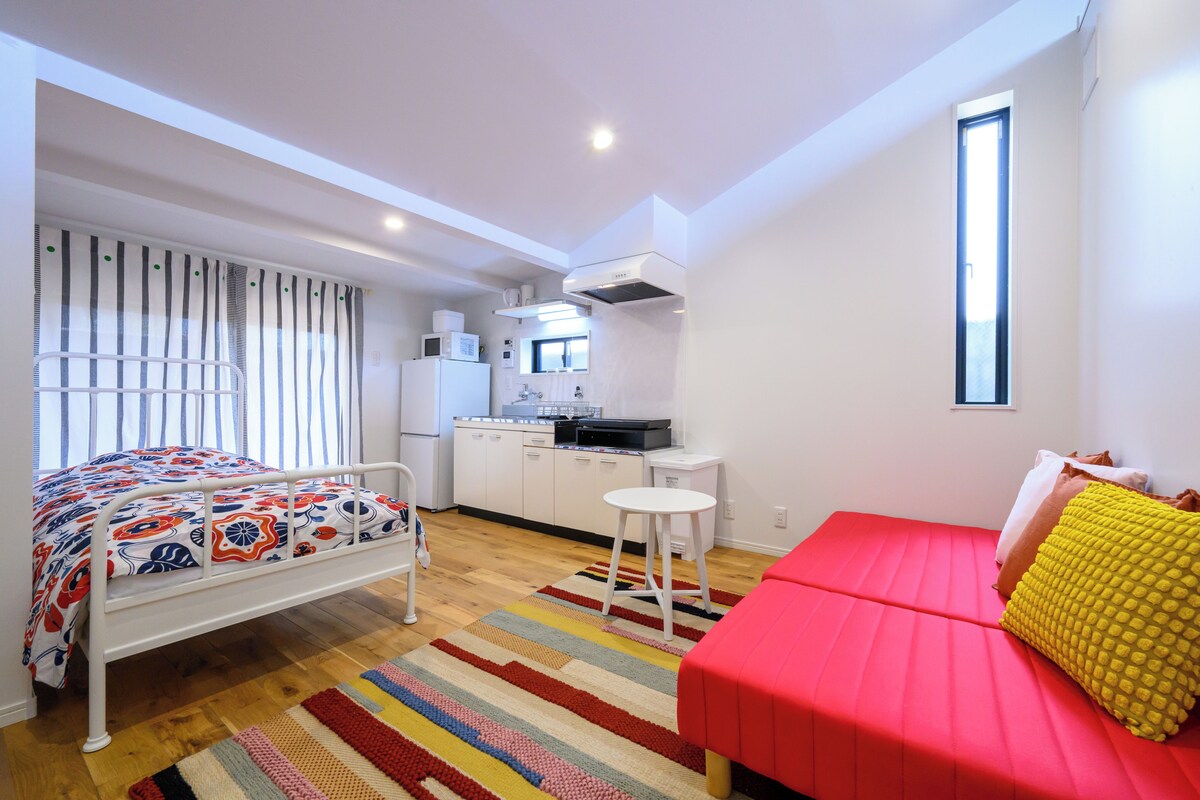 A bedroom with a bed, a red sofa, a small table, and a kitchenette in the background.