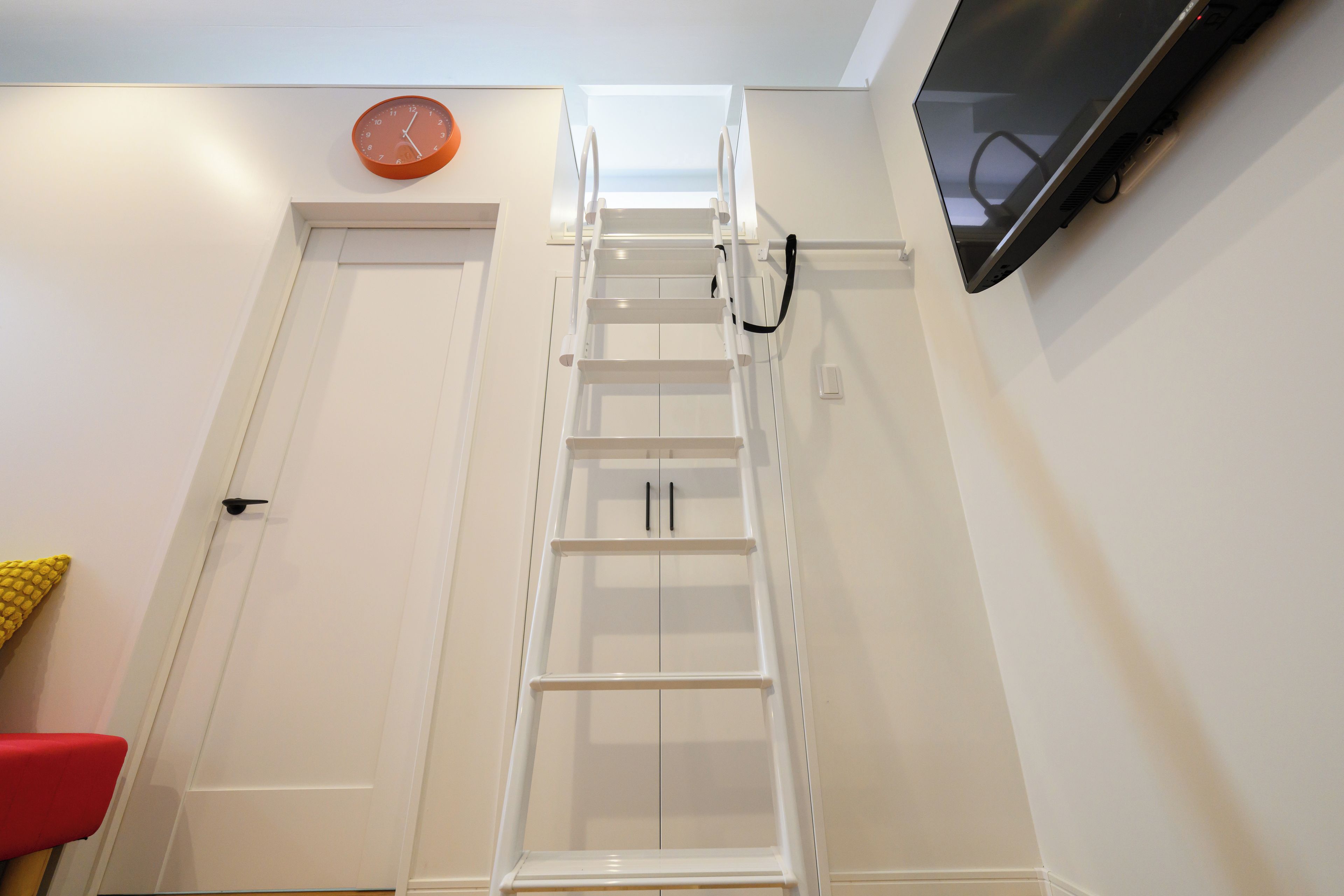 A white door with a small orange clock above it, a white ladder against the wall, and a red chair to the left.
