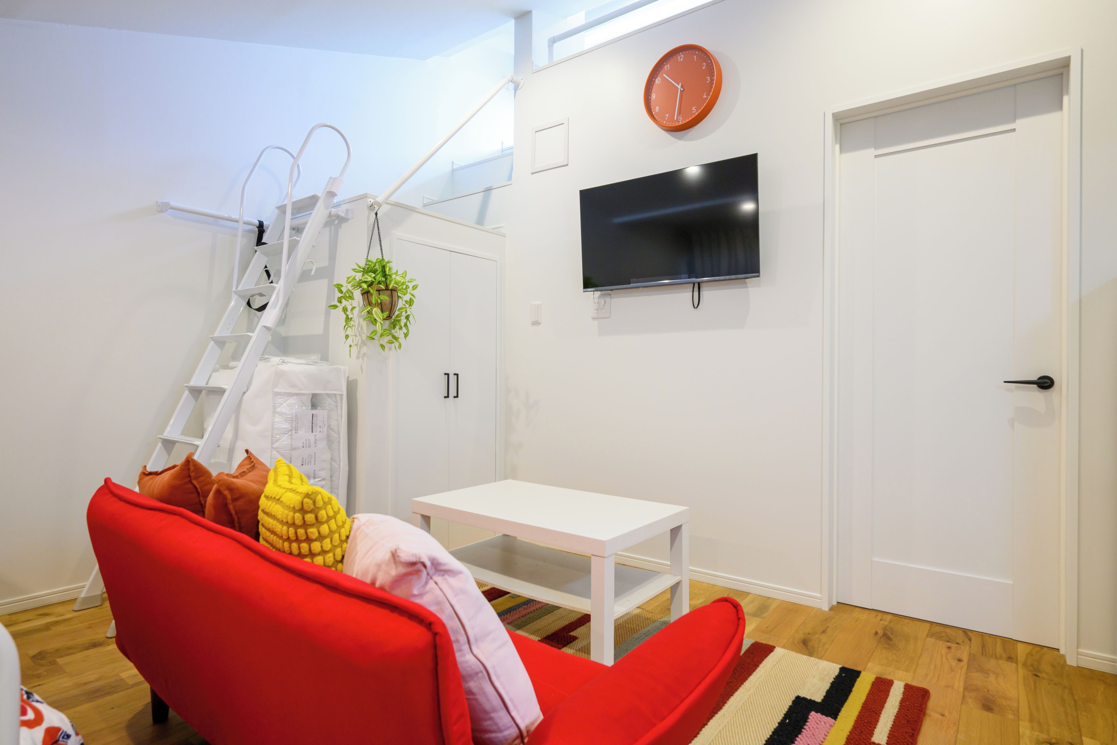 Una sala moderna con un sofá rojo, una mesa blanca, un televisor montado en la pared, una puerta blanca y una escalera con una planta.