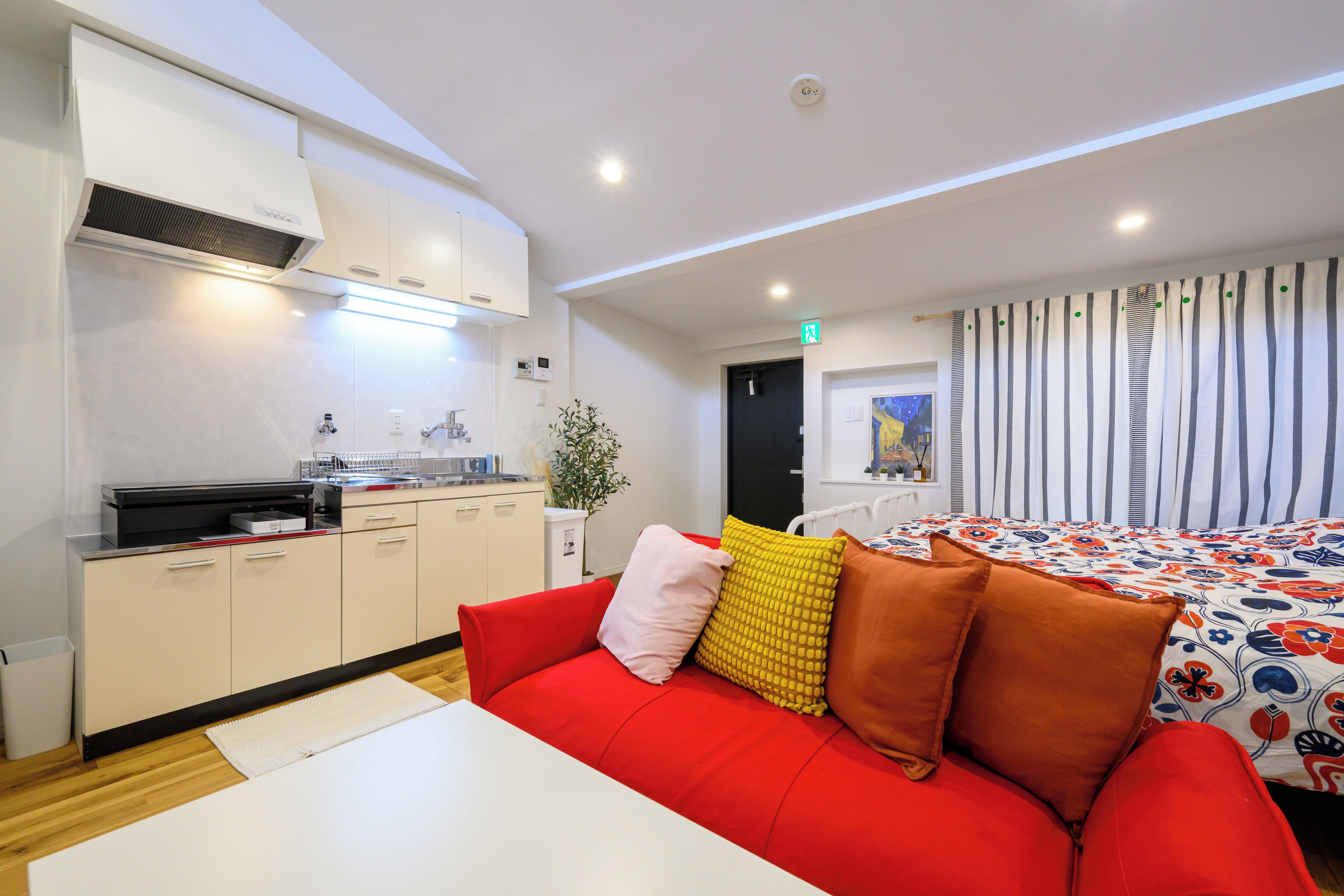 A modern hotel room with a kitchenette, a red sofa with cushions, a bed with a colorful bedspread, and a window with striped curtains.