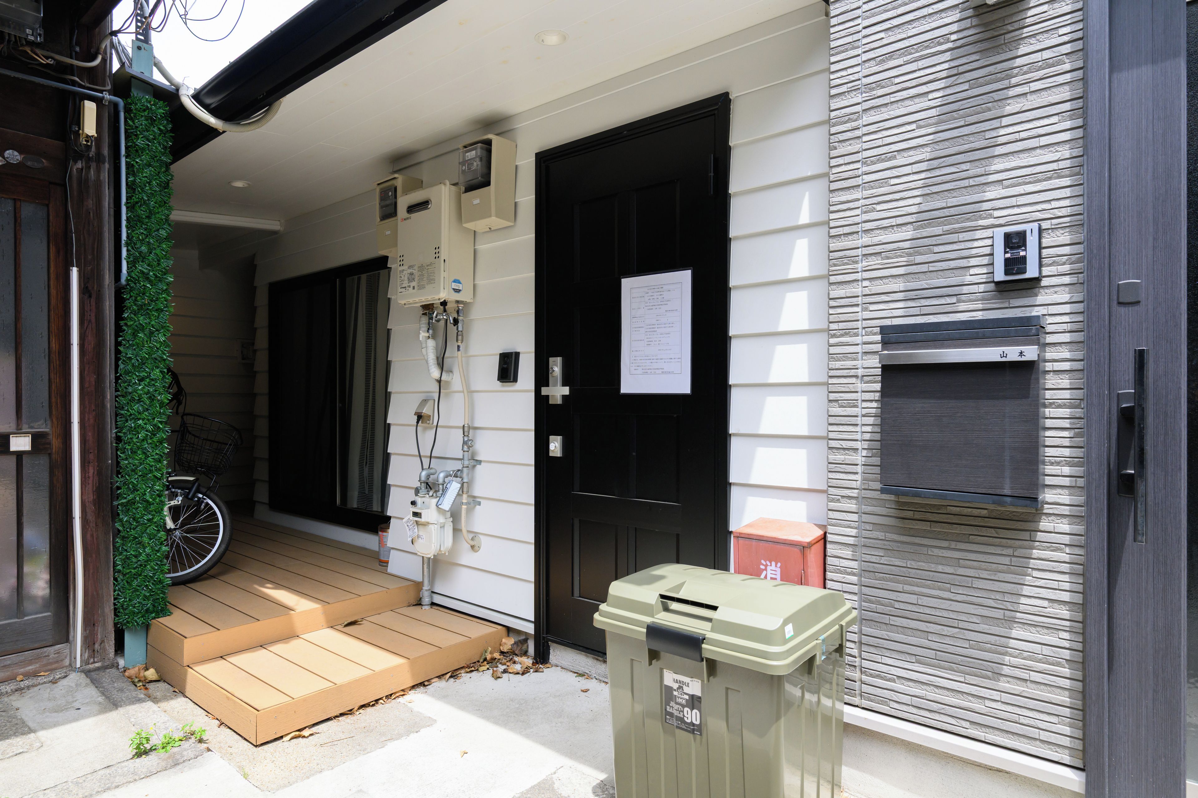 A residential entrance with a wooden door, a small step leading to a porch, a bicycle, and a trash bin.