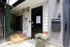 A residential entrance with a wooden door, a small step leading to a porch, a bicycle, and a trash bin. - Villa Shimabara (Kyoto)
