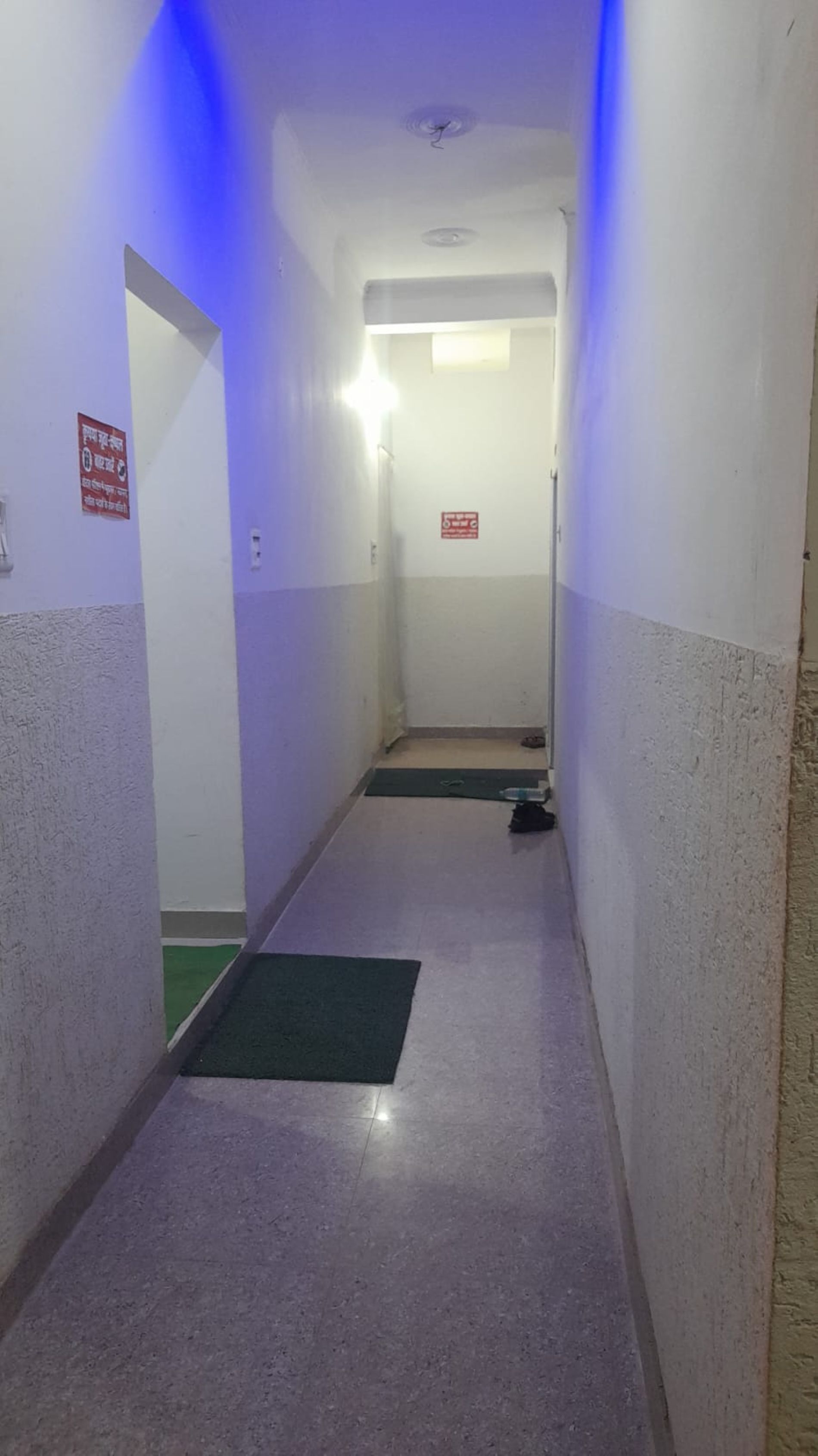 Un corridoio stretto con un tappetino verde all'ingresso, una segnaletica di uscita di sicurezza rossa e un lampadario a soffitto.