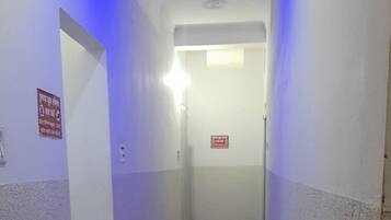 Un corridoio stretto con un tappetino verde all'ingresso, una segnaletica di uscita di sicurezza rossa e un lampadario a soffitto.