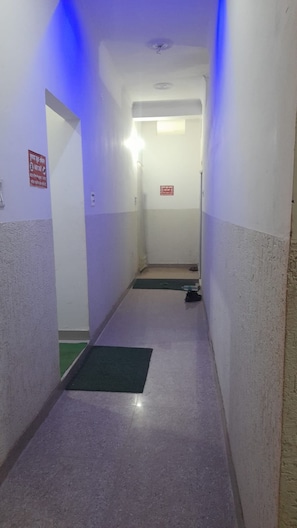 Un pasillo estrecho con un felpudo verde en la entrada, un rótulo rojo de salida de emergencia y una lámpara en el techo.