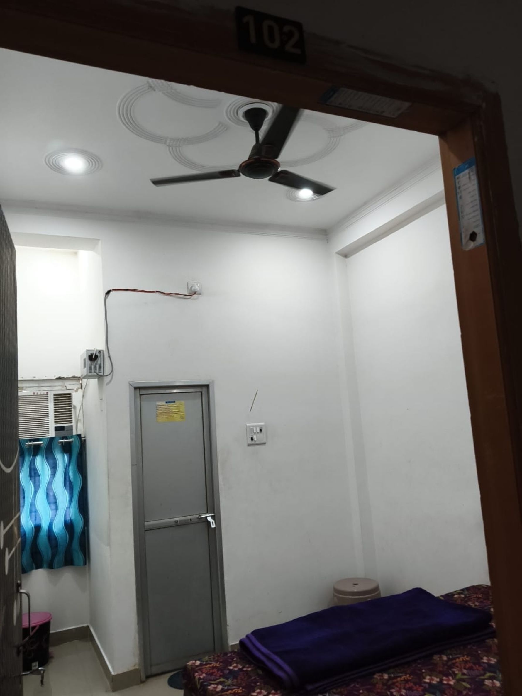Ein Zimmer mit Deckenventilator, einer Tür, einem kleinen Fenster und einem Bett mit blauer Bettdecke.