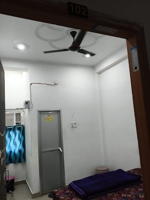 A room with a ceiling fan, a door, a small window, and a bed with a blue cover. - Maruti Nandan Hotel (Rajnagar)