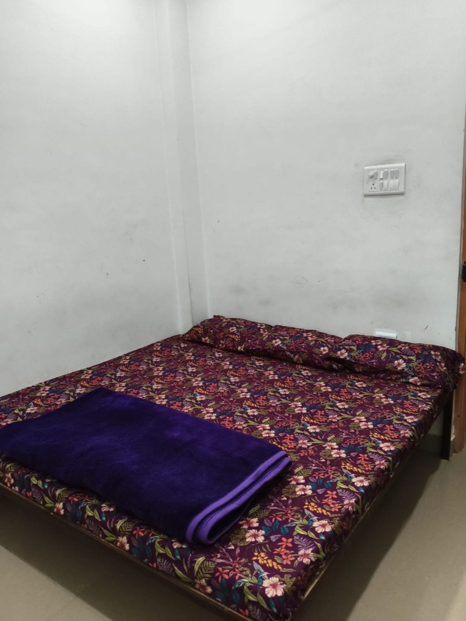 Una cama individual con una colcha floral y una manta morada en una habitación con paredes blancas y un enchufe visible.