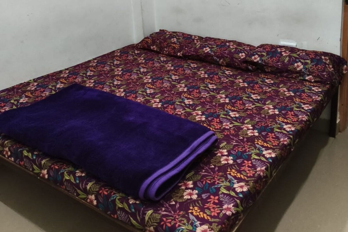 A single bed with a floral bedspread and a purple blanket in a room with white walls and a visible electrical outlet.