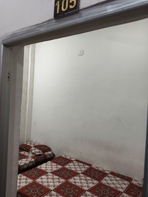 A small, enclosed room with a bed featuring a patterned bedspread. A number sign is visible above the bed. - Maruti Nandan Hotel (Rajnagar)