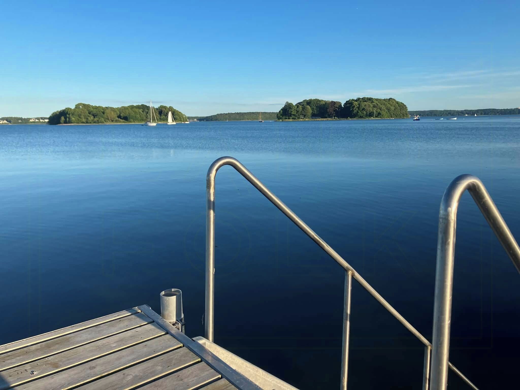 Eine Holzsteg, der sich in ruhiges Wasser erstreckt, mit Metallgeländern, umgeben von blauem Himmel und weiter Küste.