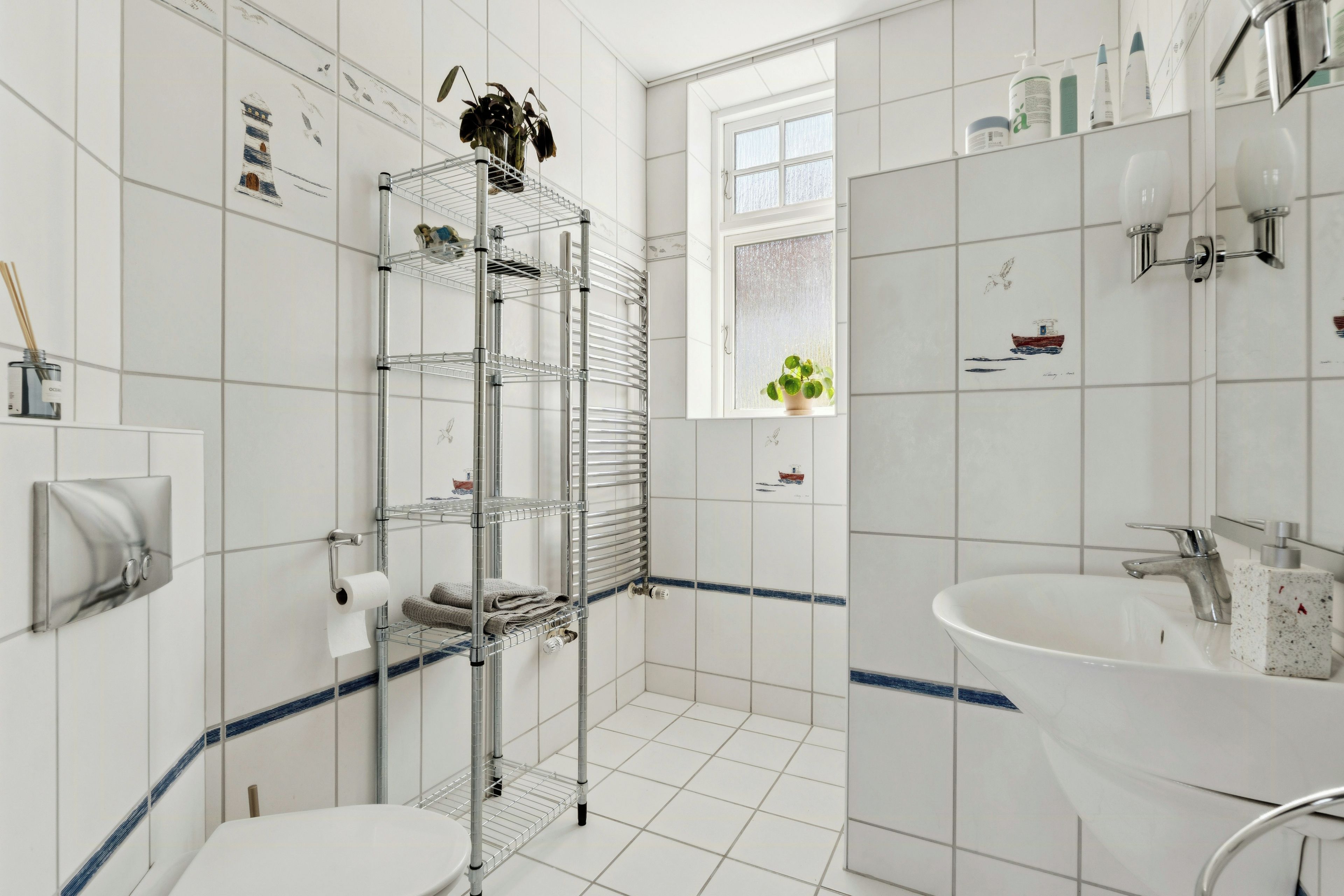A bathroom with white tiles, a metal shelving unit, a window with a view, a sink, and a toilet.