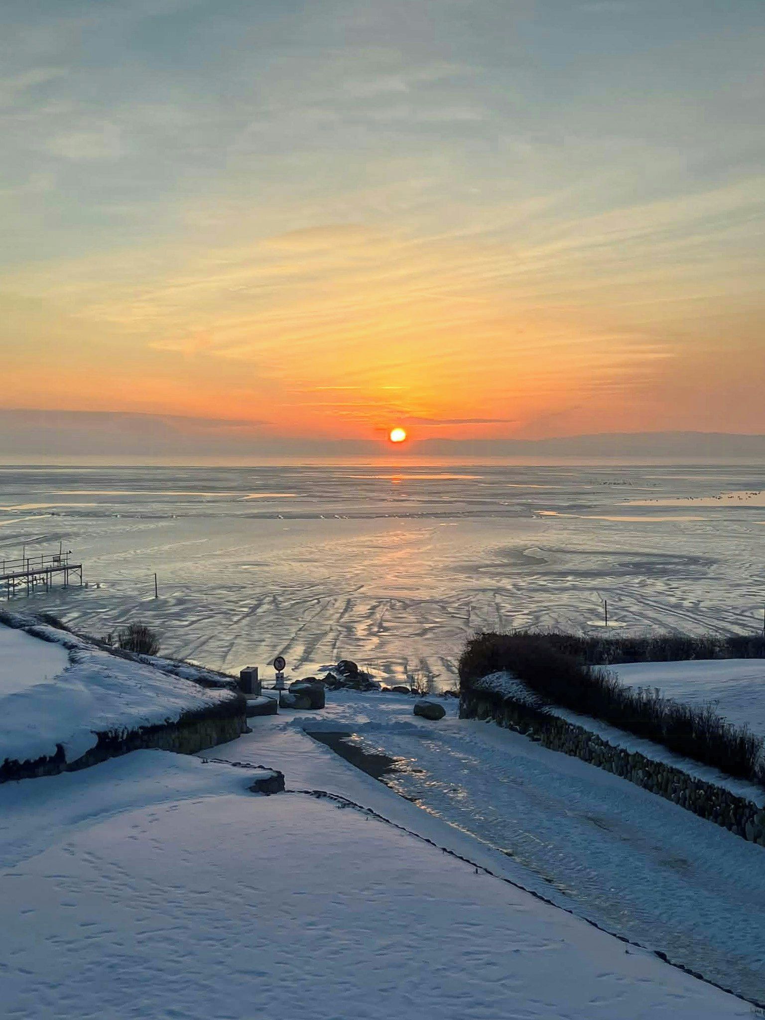 Un paysage hivernal avec un coucher de soleil sur une étendue d’eau, un sentier enneigé et une petite construction sur laquelle on peut voir une enseigne.