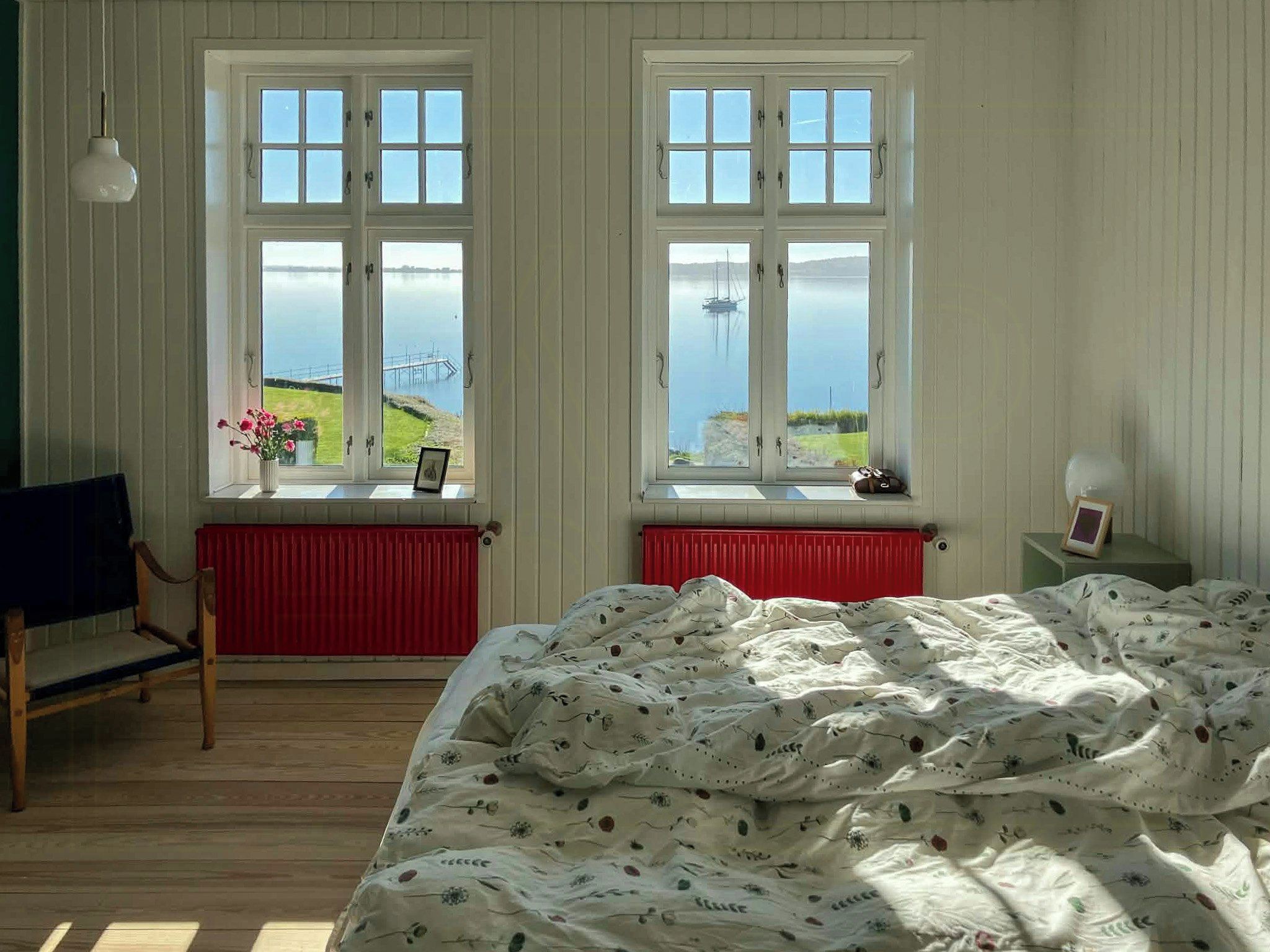 A bedroom with a bed, two windows with a view of the sea, a chair, and a radiator.