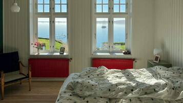A bedroom with a bed, two windows with a view of the sea, a chair, and a radiator.