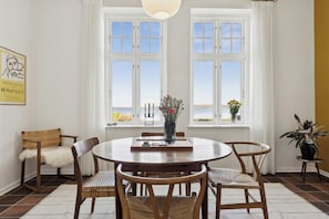 Een eetruimte met een ronde tafel, stoelen en uitzicht op zee door grote ramen.