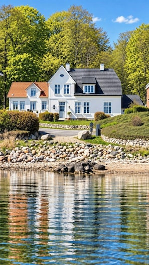 Een groot wit huis met een rood dak, gelegen naast een waterpartij met een rotsachtige oever.