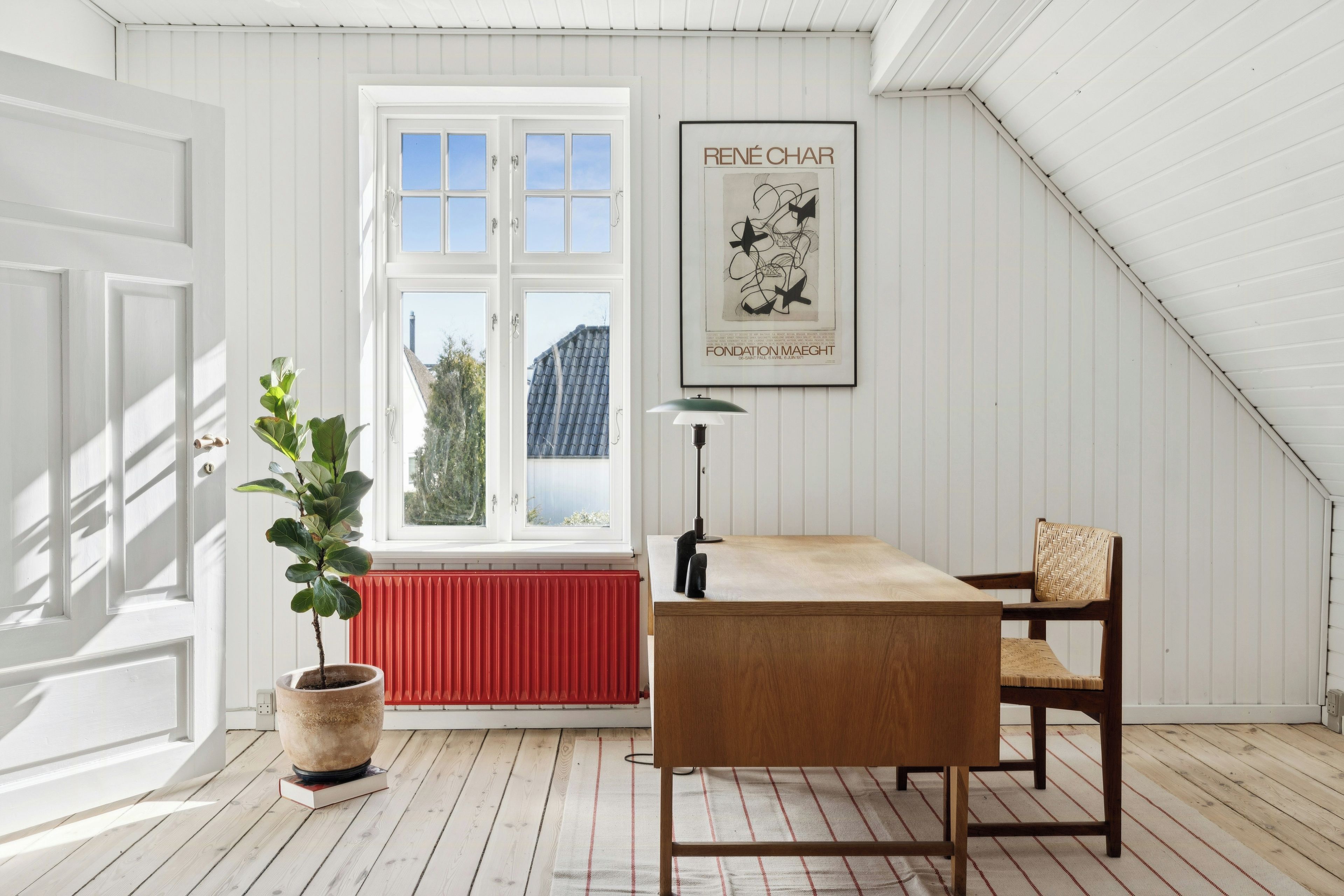Een kamer met een witte deur, een raam met uitzicht, een kunstwerk in een lijst aan de muur, een bloempot, een tafel en een stoel.