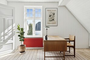 Una habitación con puerta blanca, ventana con vistas, una obra de arte enmarcada en la pared, planta en maceta, una mesa y una silla.