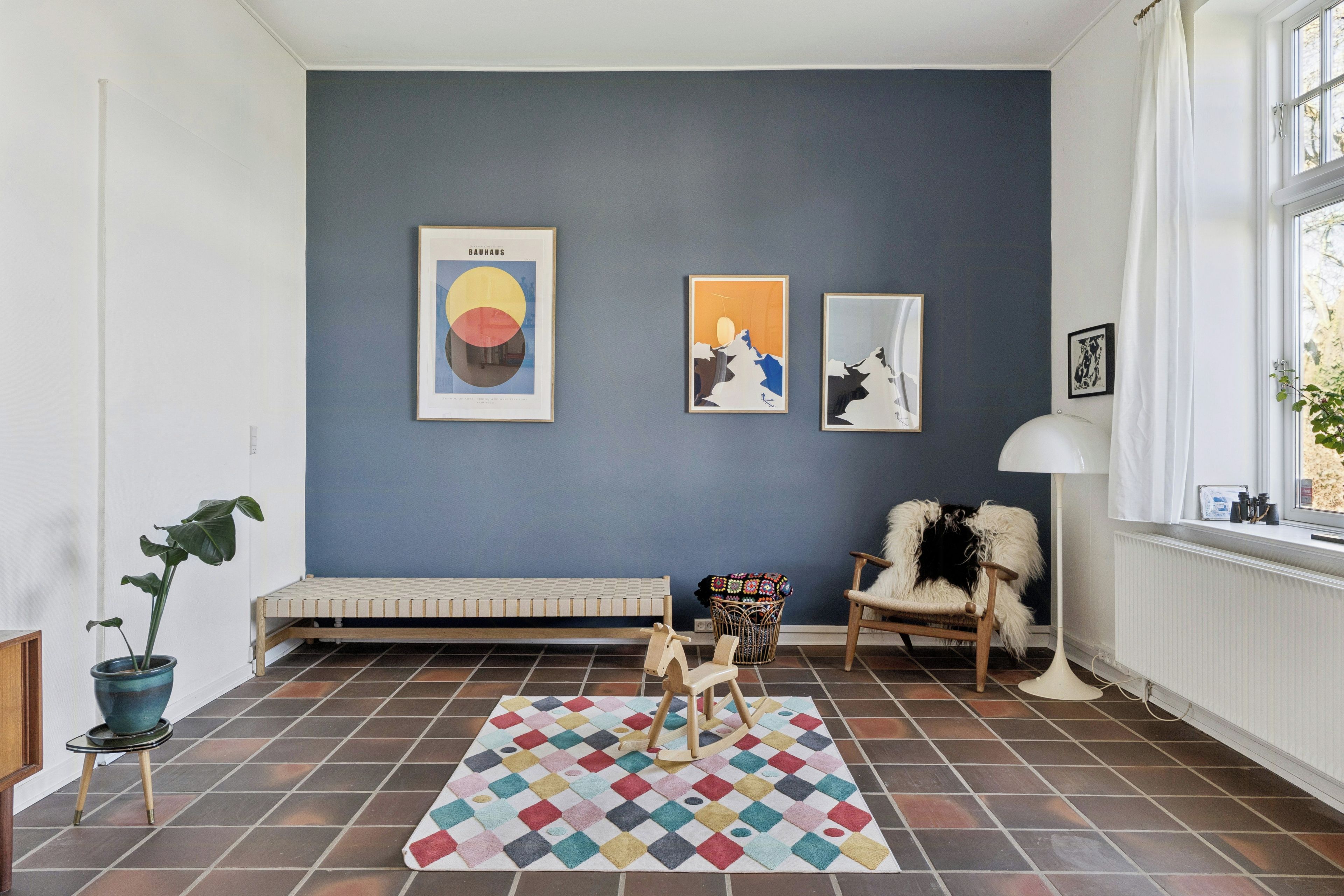 Een kamer met een blauwe wand, een bankje, een schommelstoel, een kleurrijk vloerkleed en kunstwerken in lijsten.