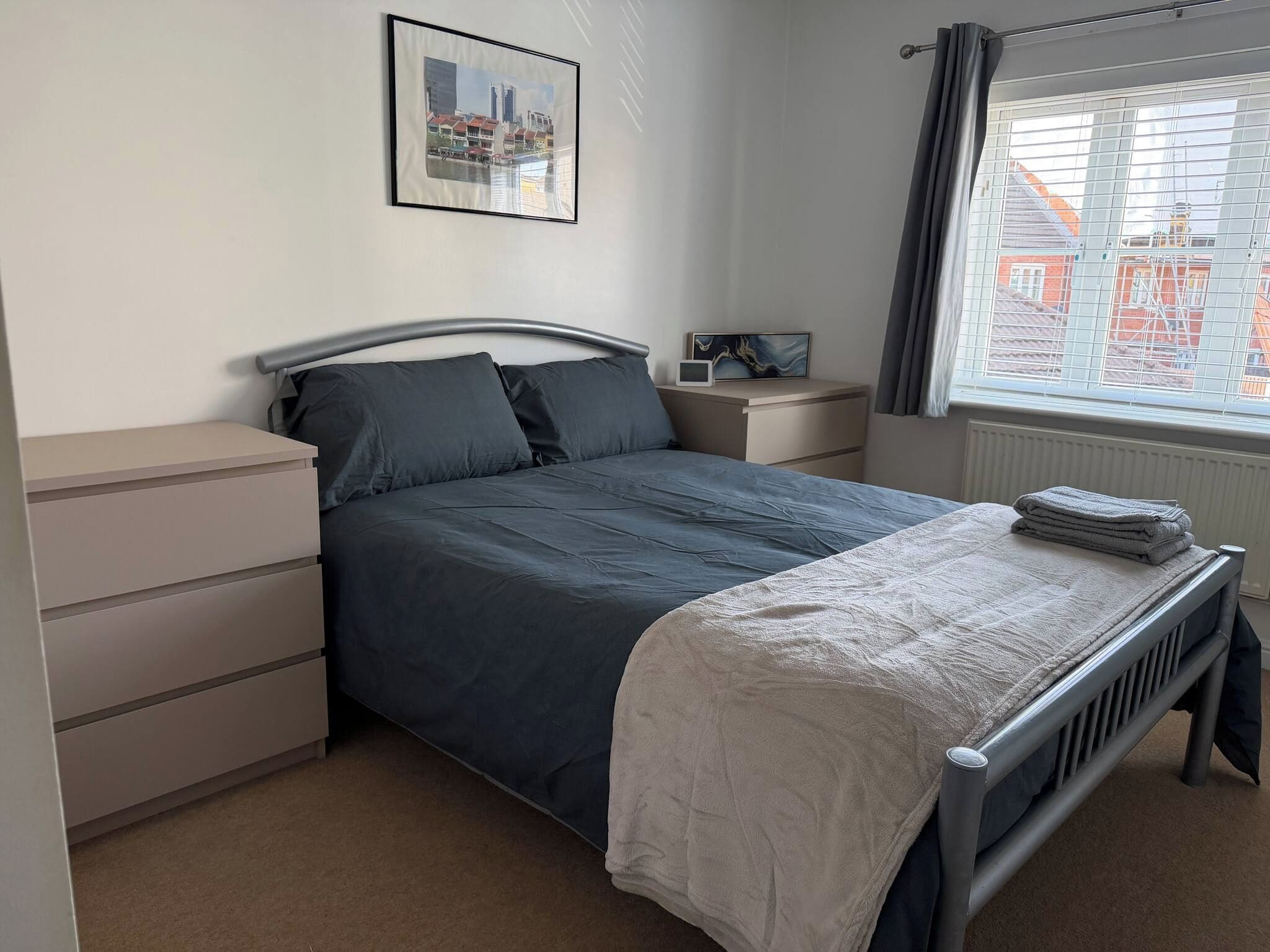 Quarto com cama, mesa de cabeceira, janela com cortinas e um quadro na parede.