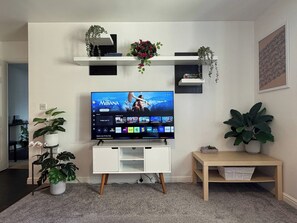 A living room with a flat-screen TV mounted on a white stand, surrounded by potted plants and a wooden side table. - A&R Guest House - 2-bed Coach House, Bristol (Bristol)
