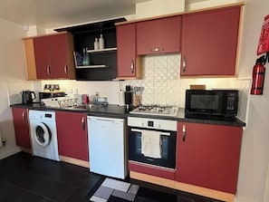 A compact kitchen with red cabinets, a washing machine, a dishwasher, a microwave, and a gas stove. - A&R Guest House - 2-bed Coach House, Bristol (Bristol)