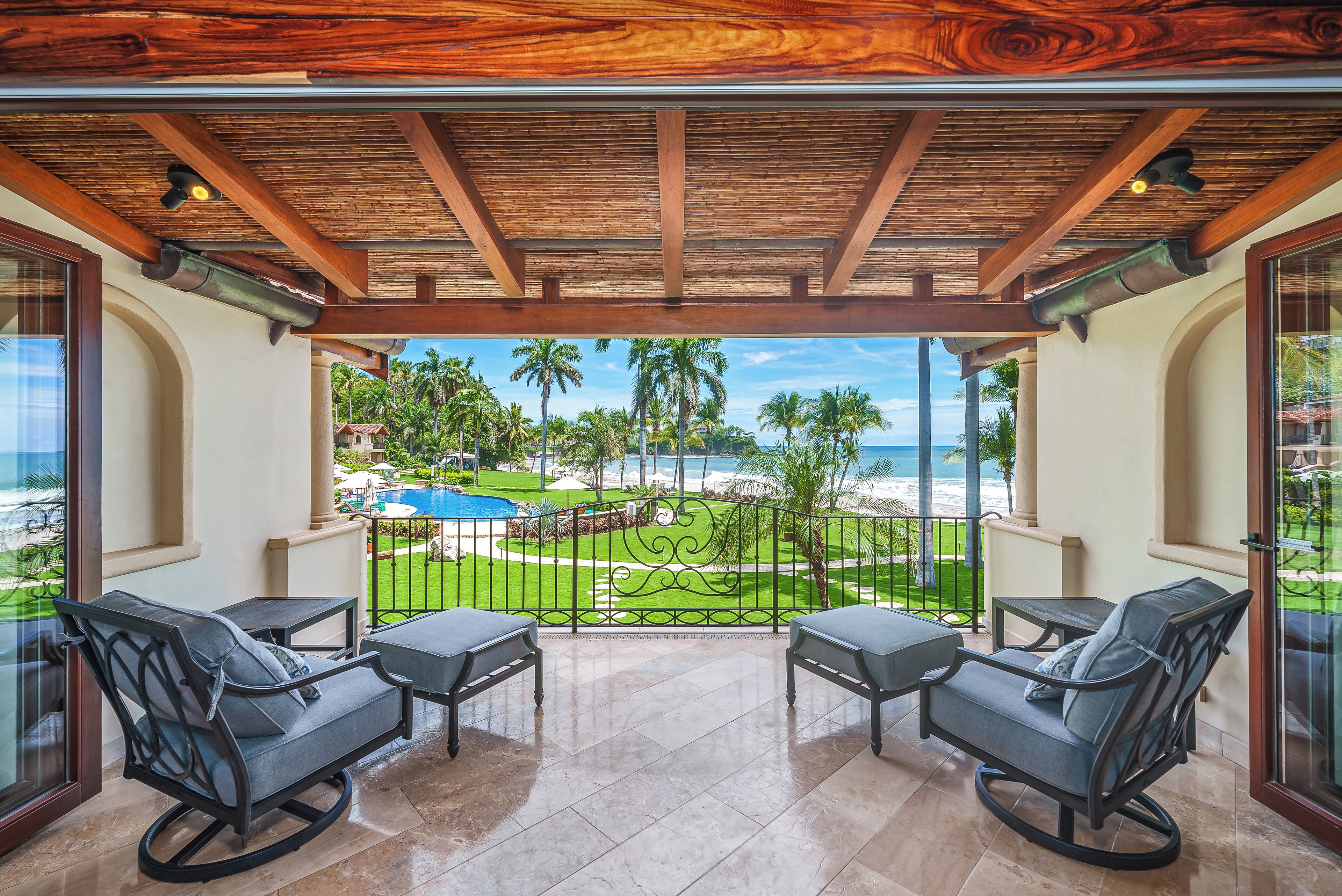 A balcony with two chairs and a table, overlooking a pool and palm trees.