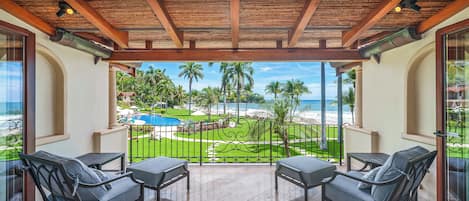 A balcony with two chairs and a table, overlooking a pool and palm trees.