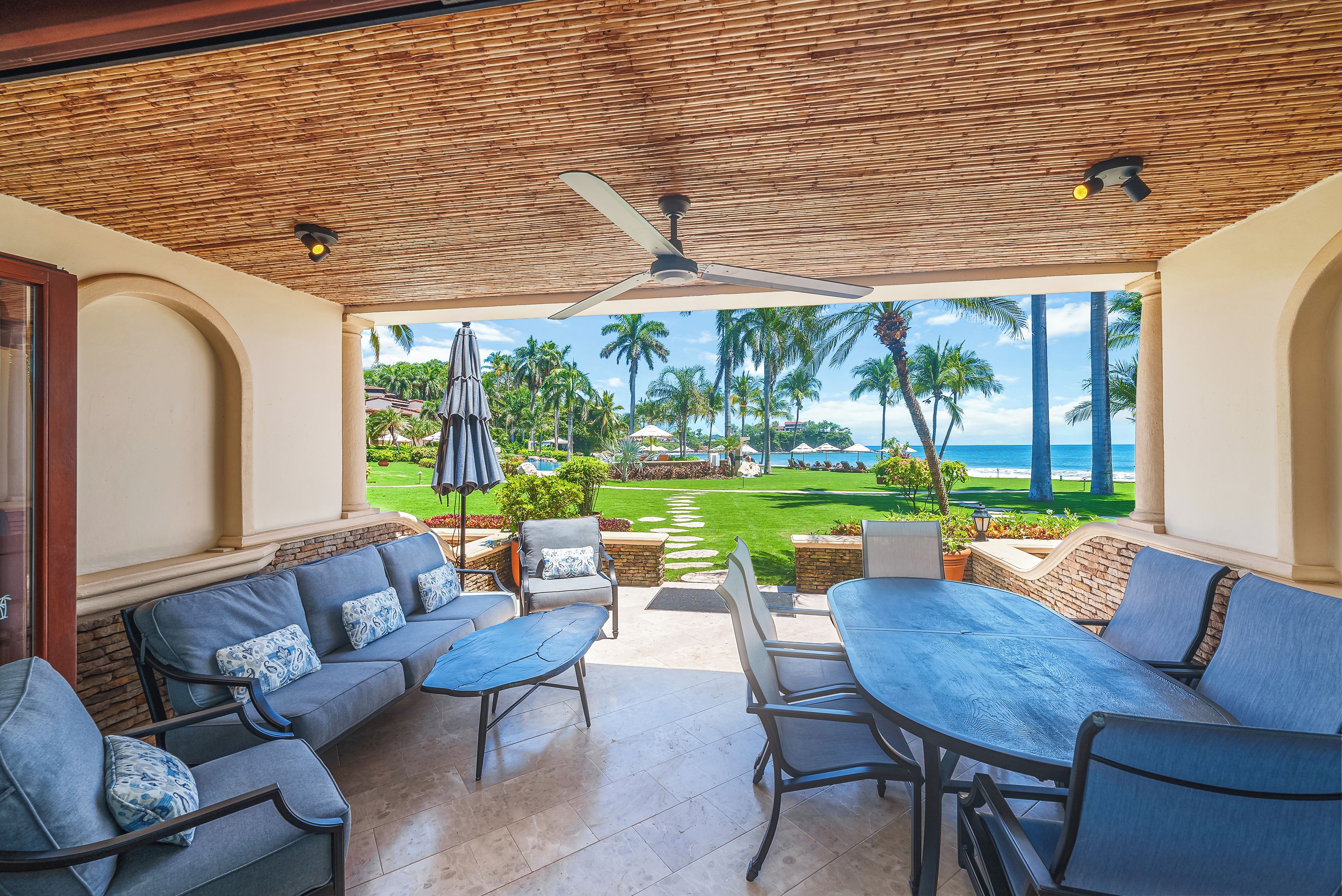 A covered outdoor seating area with a view of the ocean and palm trees.