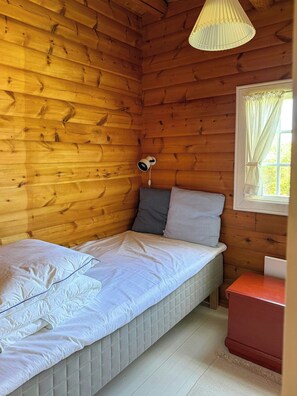 Ein kleines Zimmer mit einem Einzelbett, Holzboden, einer Nachttischlampe, einem Fenster mit Vorhängen und einem kleinen roten Tisch.