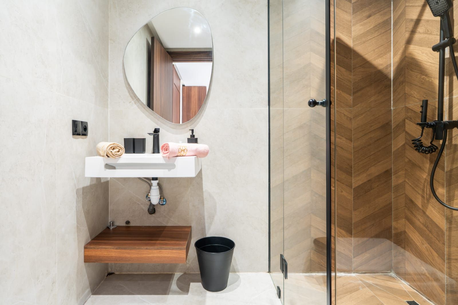 A modern bathroom with a glass shower enclosure, a round mirror, a white sink with a wooden shelf, and a black trash bin.